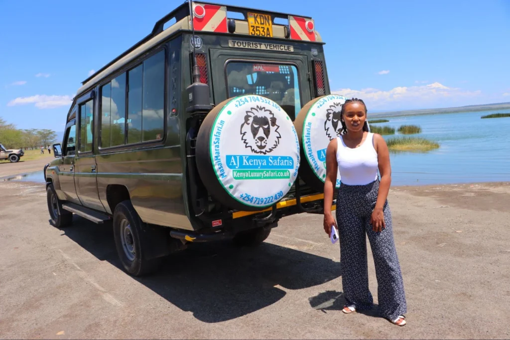A woman standing next to a 4x4 safari vehicle parked by a lake.