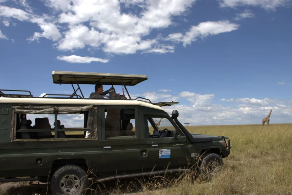 Tourists standing in an open-roof safari Land Cruiser watching a giraffe on the savanna.