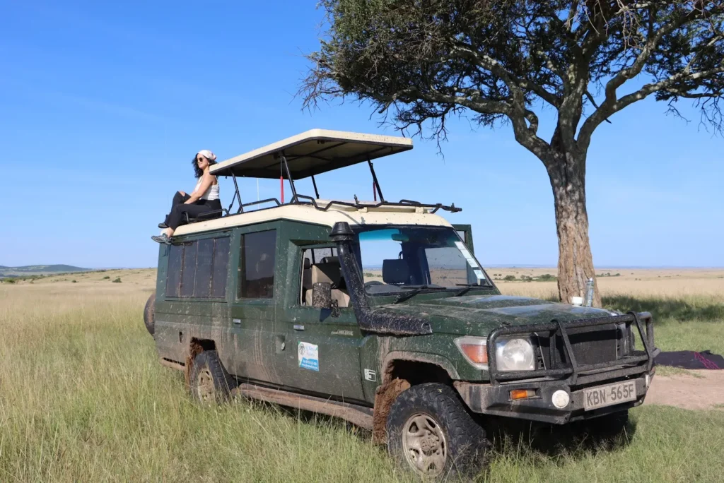 Woman sitting on a safari Land Cruiser roof rack next to an acacia tree.