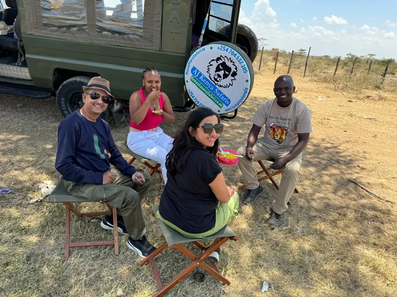 Group of tourists and their guide eating a picnic lunch beside a safari vehicle.