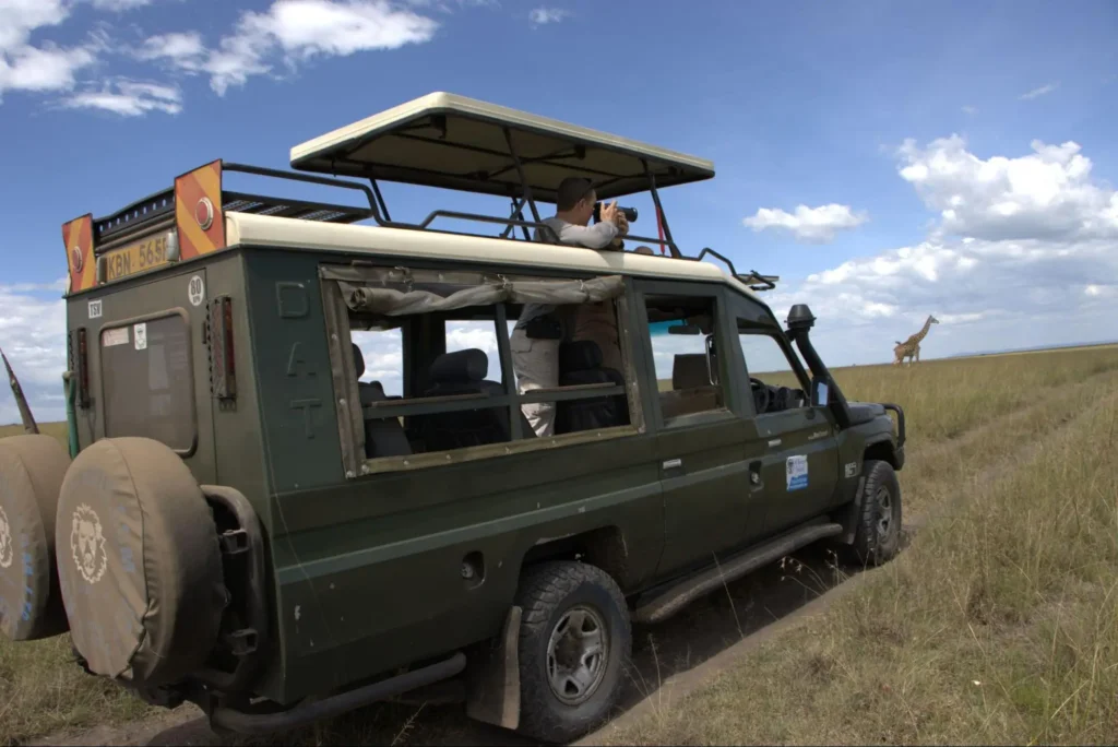 A green safari vehicle with a tourist using binoculars looking at a distant giraffe.