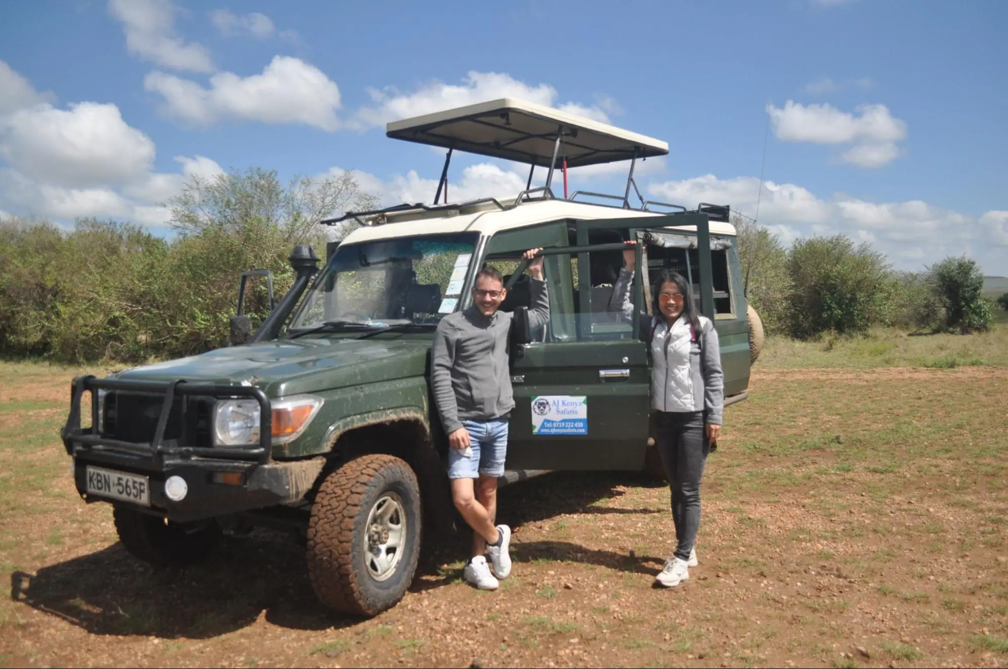 Two smiling tourists posing next to an open-roof 4x4 safari vehicle on the savanna.