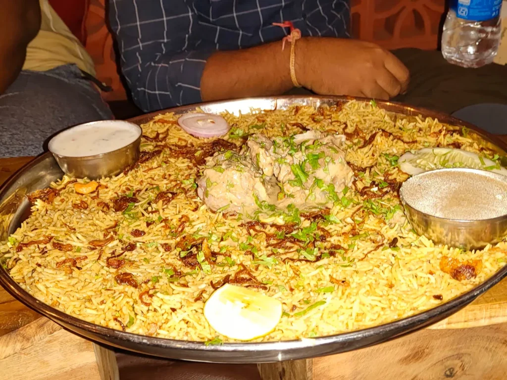 A large metal platter filled with spiced biryani rice, meat curry, and yogurt sauce.