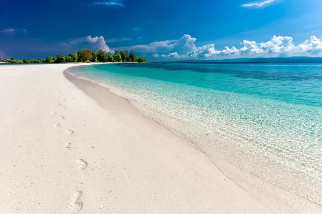 Footprints on a wide white sand beach next to calm turquoise ocean water.