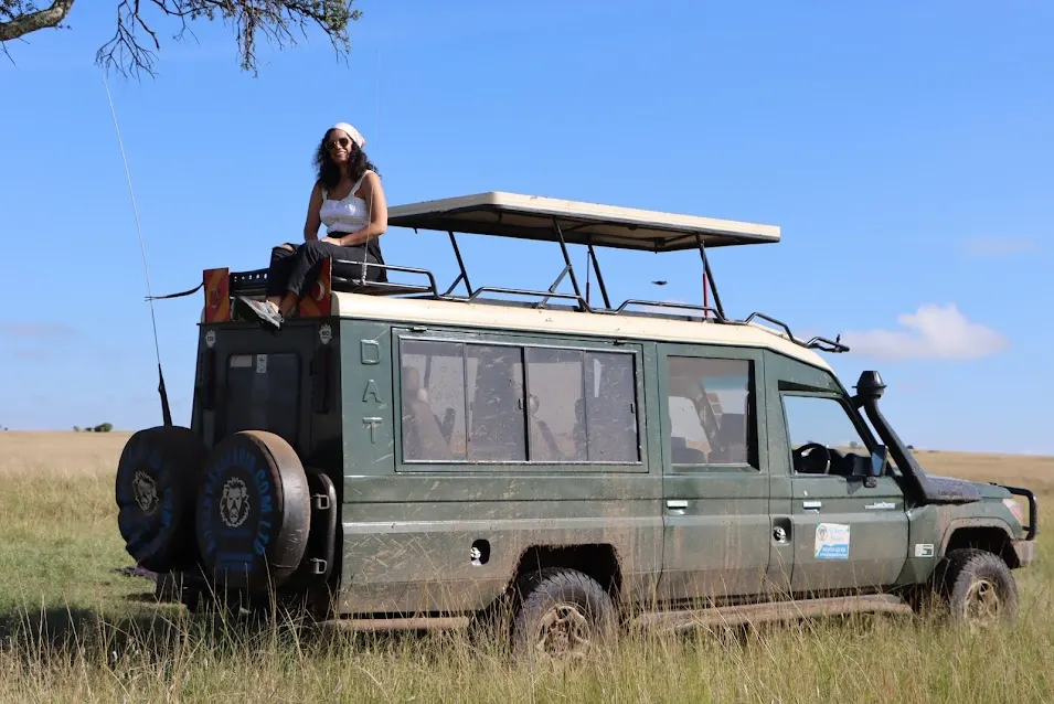 A woman sitting on the roof of a muddy green safari vehicle in open grassland.