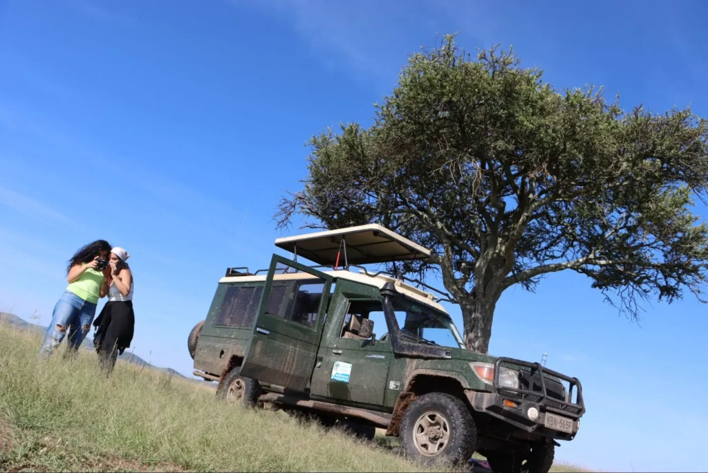 Two tourists checking photos next to a private 4x4 safari vehicle under a tree.