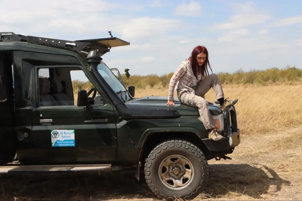 A woman in cargo pants sitting on a 4x4 safari vehicle in dry grassland.