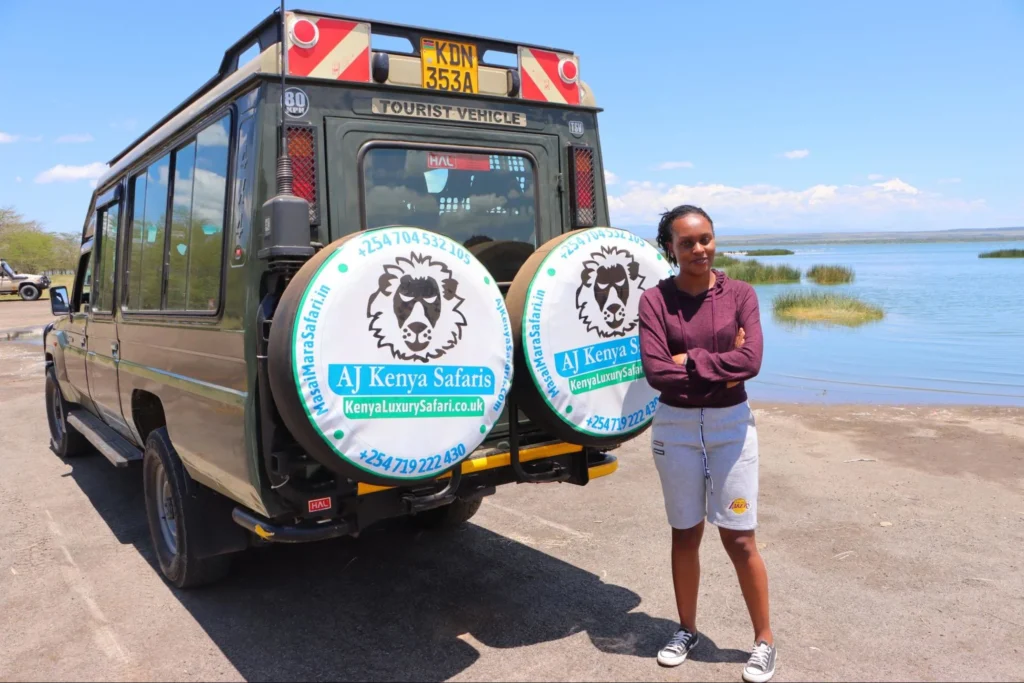 A woman standing with arms crossed next to a green safari vehicle by a lake.