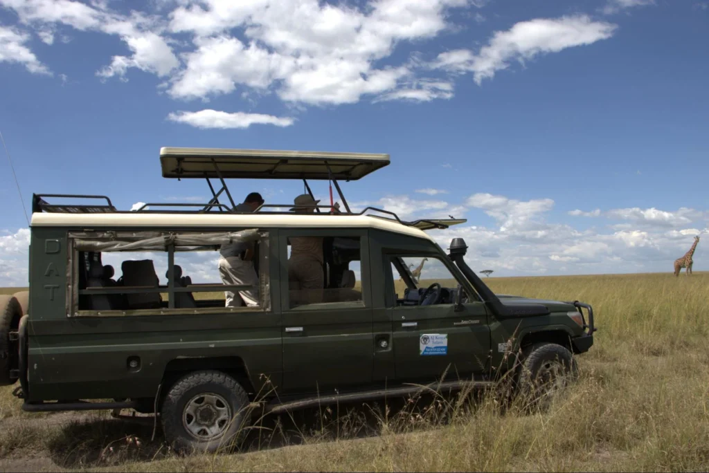 Tourists inside an open roof 4x4 safari vehicle observing a giraffe on the savanna.