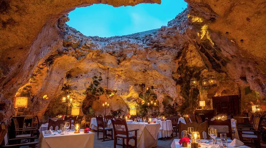 Candlelit dining tables inside a natural stone cave restaurant with an open sky ceiling.