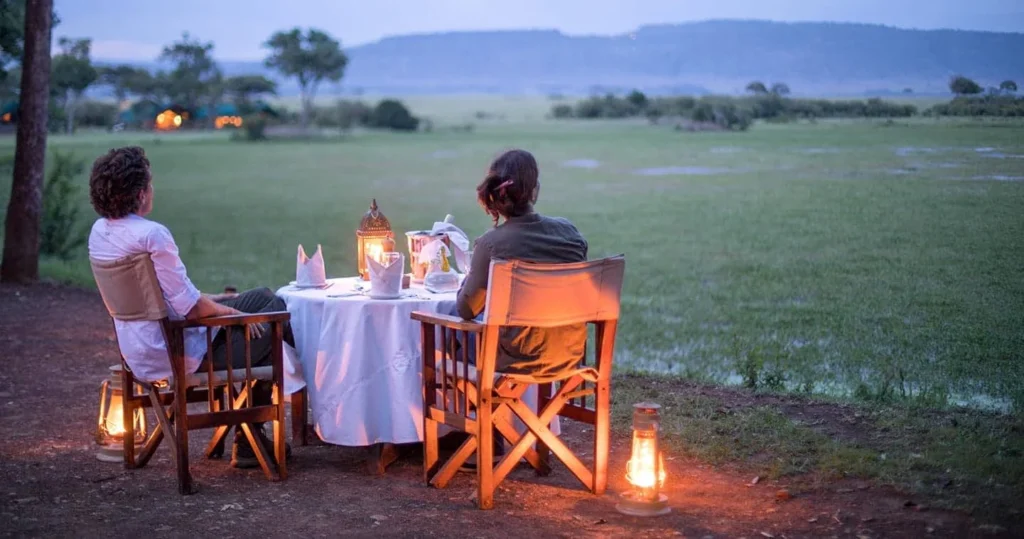 A couple enjoying a lantern-lit outdoor dinner overlooking a green marshy safari plain.
