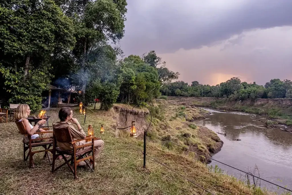Two guests relax in chairs on a high cliff edge overlooking the Mara River at dusk.