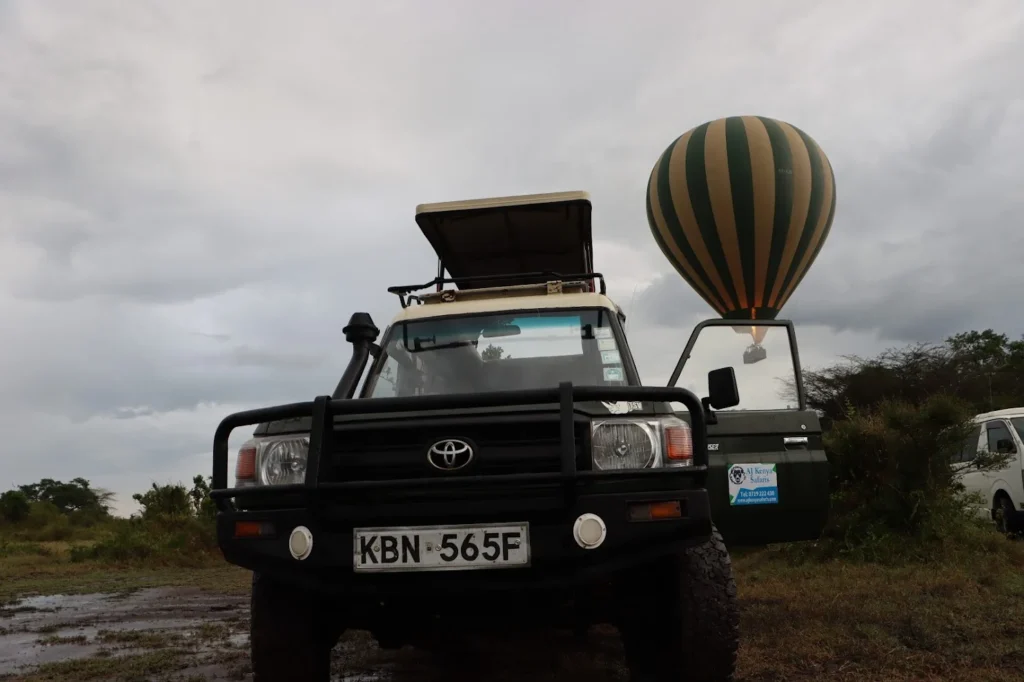 Green safari vehicle with an open roof parked beneath a striped hot air balloon.